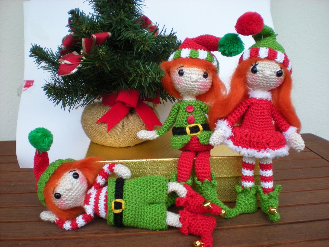 Drei festliche Amigurumi-Wichtel mit orangefarbenem Haar, rot-grünen Kleidern und Bommelmützen neben einem kleinen geschmückten Tannenbaum