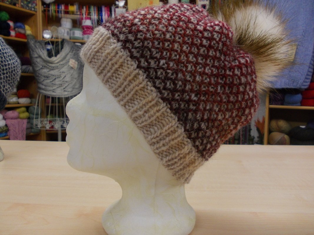 Seitliche Ansicht einer gestrickten Mütze mit Umschlagbündchen und Felloptik-Bommel auf Mannequinkopf