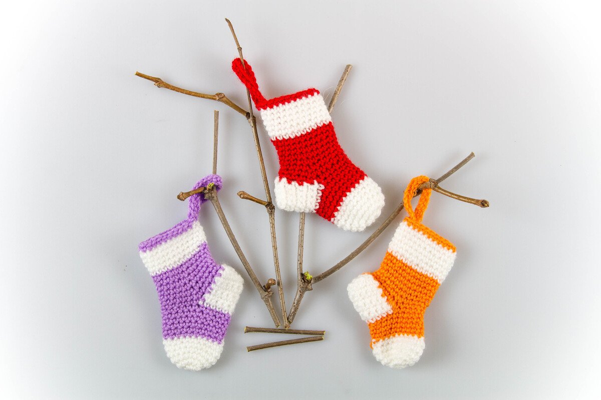 Drei gehäkelte kleine Weihnachtsstrümpfe in Rot, Lila und Orange mit weißen Abschlüssen an einem Zweig