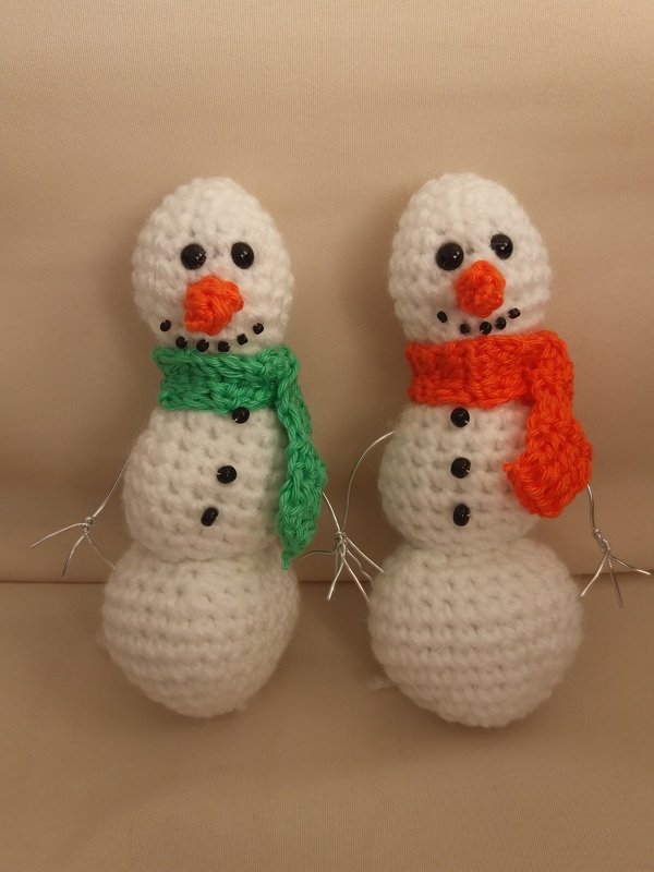 Zwei gehäkelte Amigurumi-Schneemänner mit grünem und orangefarbenem Schal, Perlenaugen und Drahtarmen