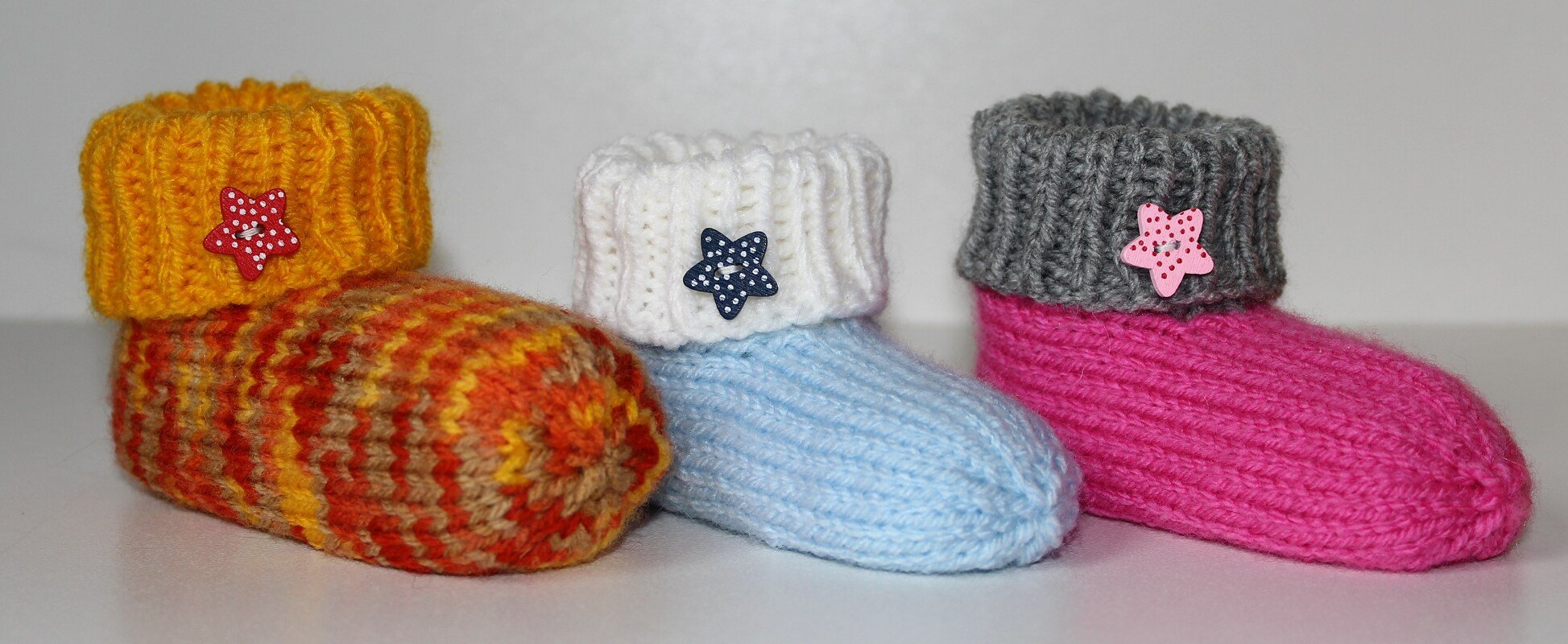 Drei gestrickte Babyschuhe: orange-gelb gestreift mit gelbem Bündchen, hellblau mit weißem Bündchen und pink mit grauem Bündchen, alle mit sternförmigen Knöpfen