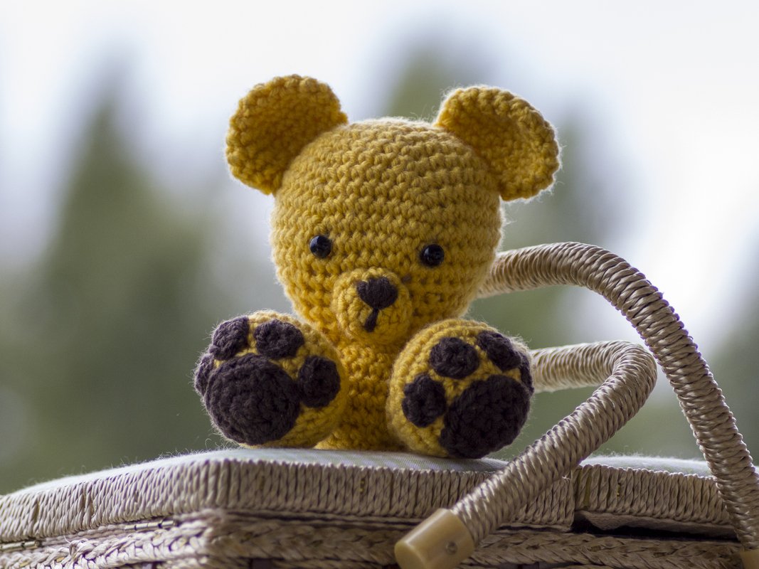 Kleiner gelber gehäkelter Amigurumi-Teddybär mit dunkelbraunen Tatzen auf einem geflochtenen Korb