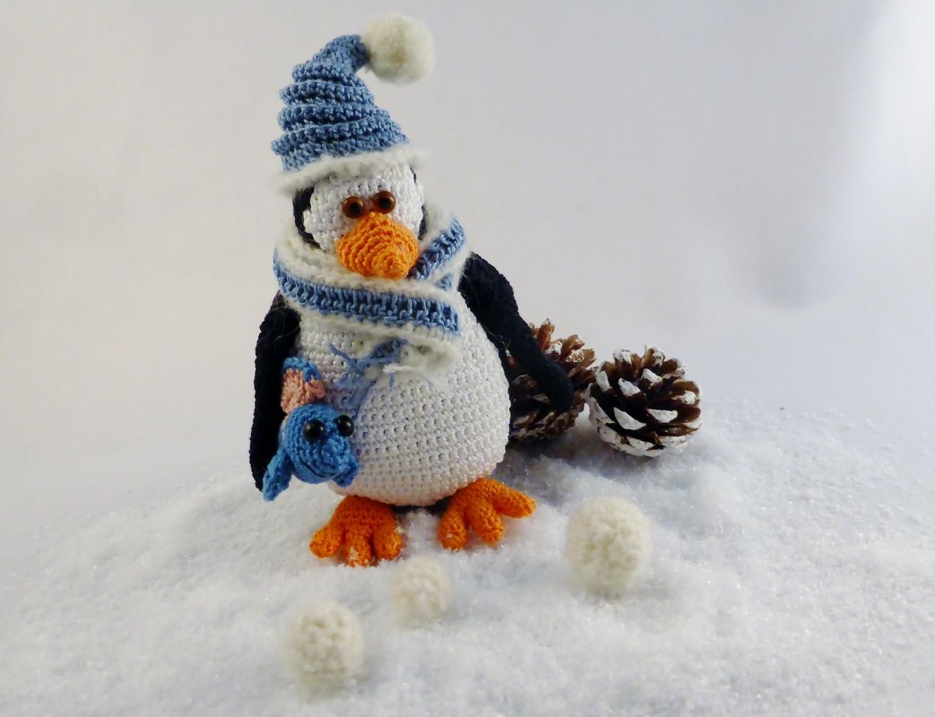 Gehäkelter Pinguin mit Mütze, Schal und kleinem blauen Vogel in einer Winterlandschaft mit Zapfen und gehäkelten Schneebällen.