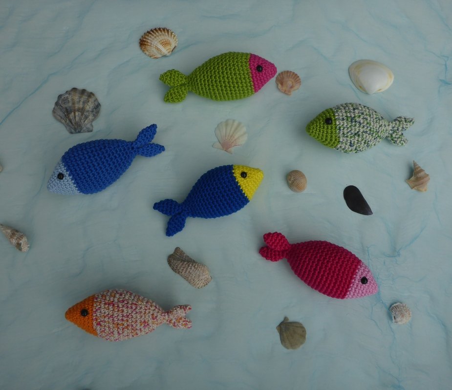 Sieben gehäkelte Amigurumi-Fische in verschiedenen Farben mit Muscheln auf hellem Stoff