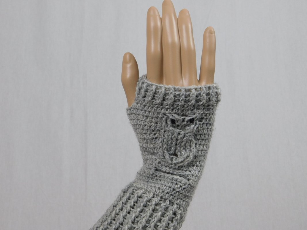Mannequin-Hand mit hellgrauem, gehäkeltem fingerlosen Handschuh und Eulenrelief auf dem Handrücken.