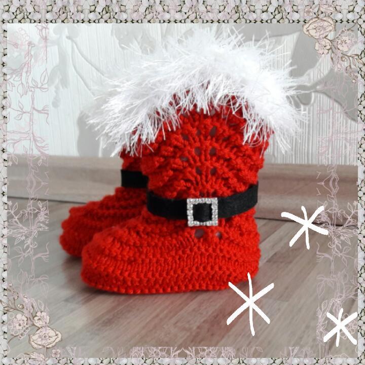 Zwei rote, gestrickte Babyschuhe im Weihnachtsmann-Look mit weißem Plüschrand und schwarzem Gürtel mit Zierschnalle auf Holzuntergrund.