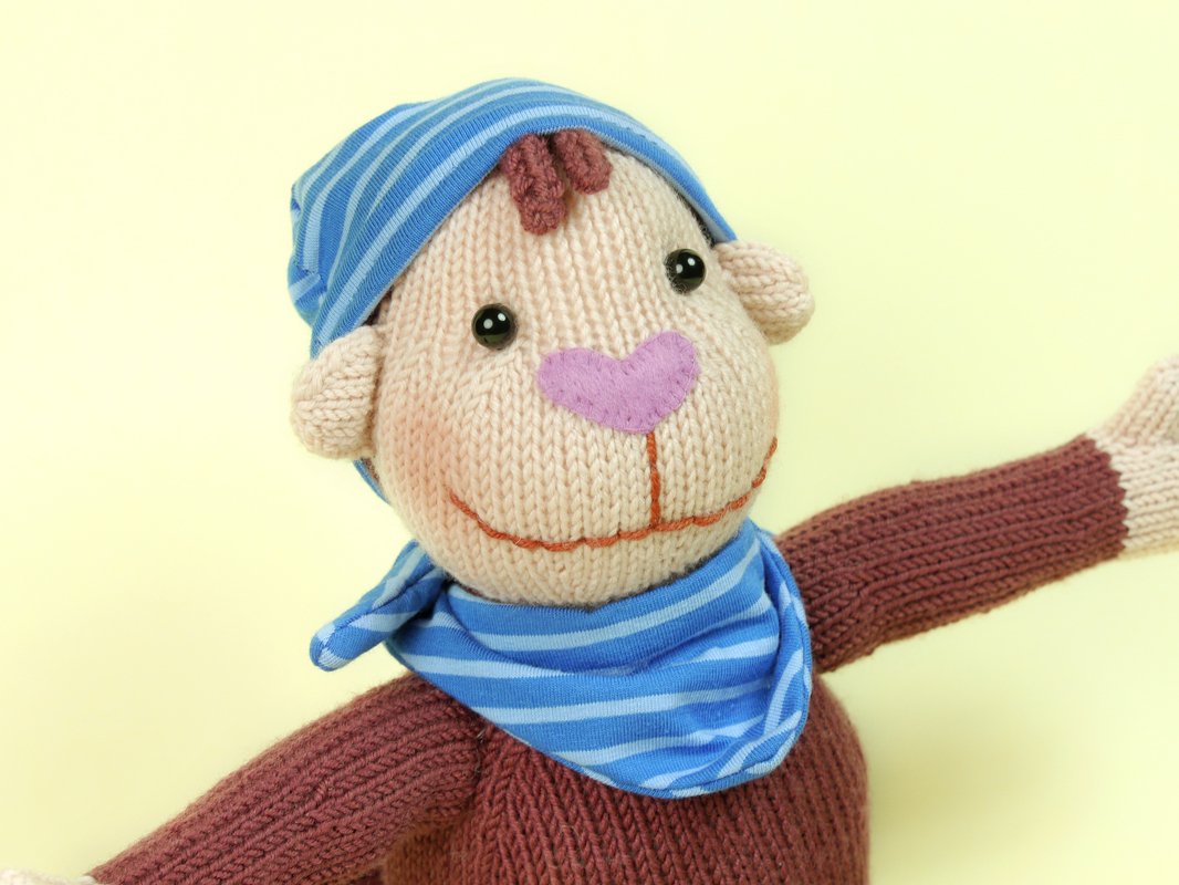 Knitted amigurumi monkey with beige face, brown body, blue striped hat and scarf on a pale yellow background.