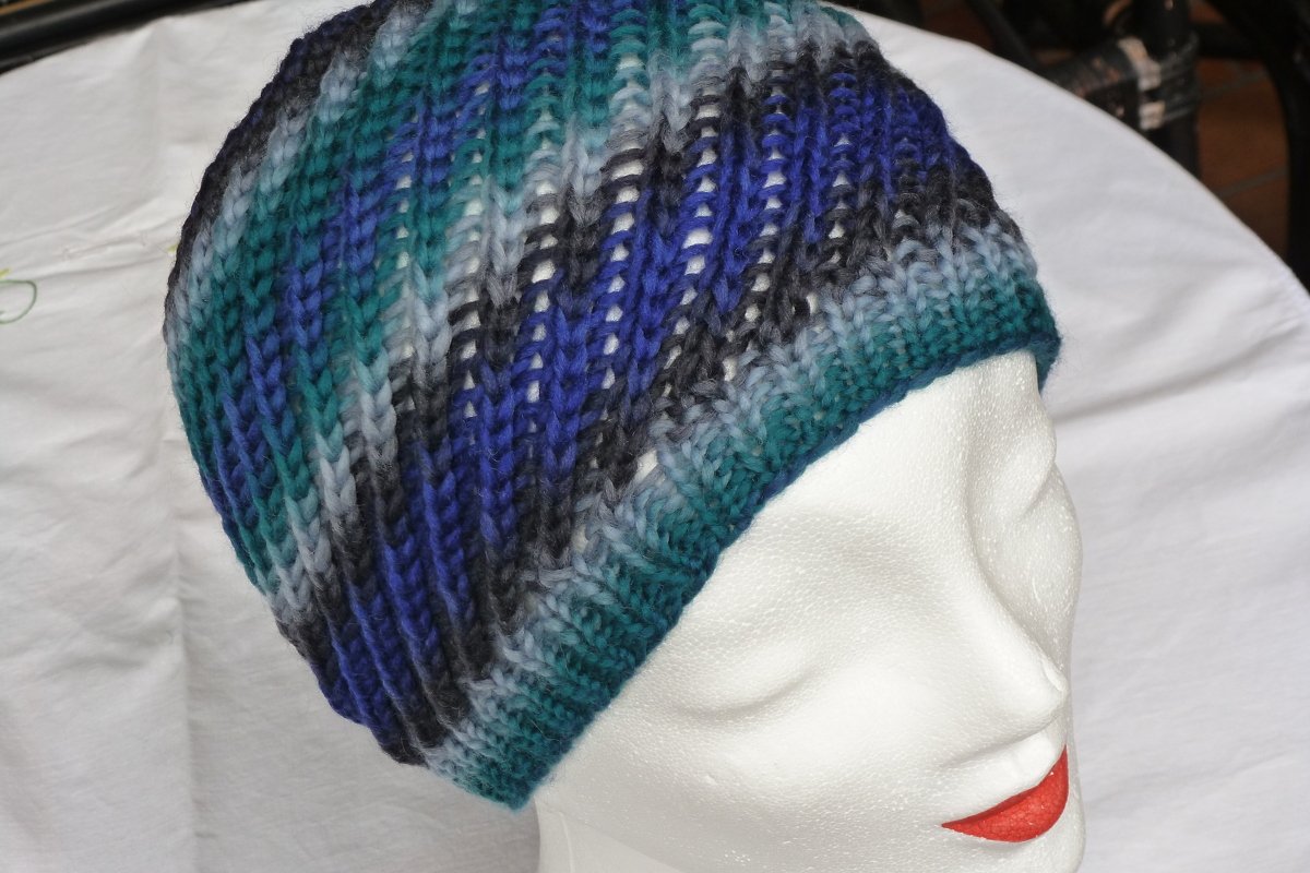Gestrickte Beanie-Mütze in Blau-, Türkis- und Grautönen auf weißem Schaufensterkopf. Vertikales Rippenmuster erkennbar.