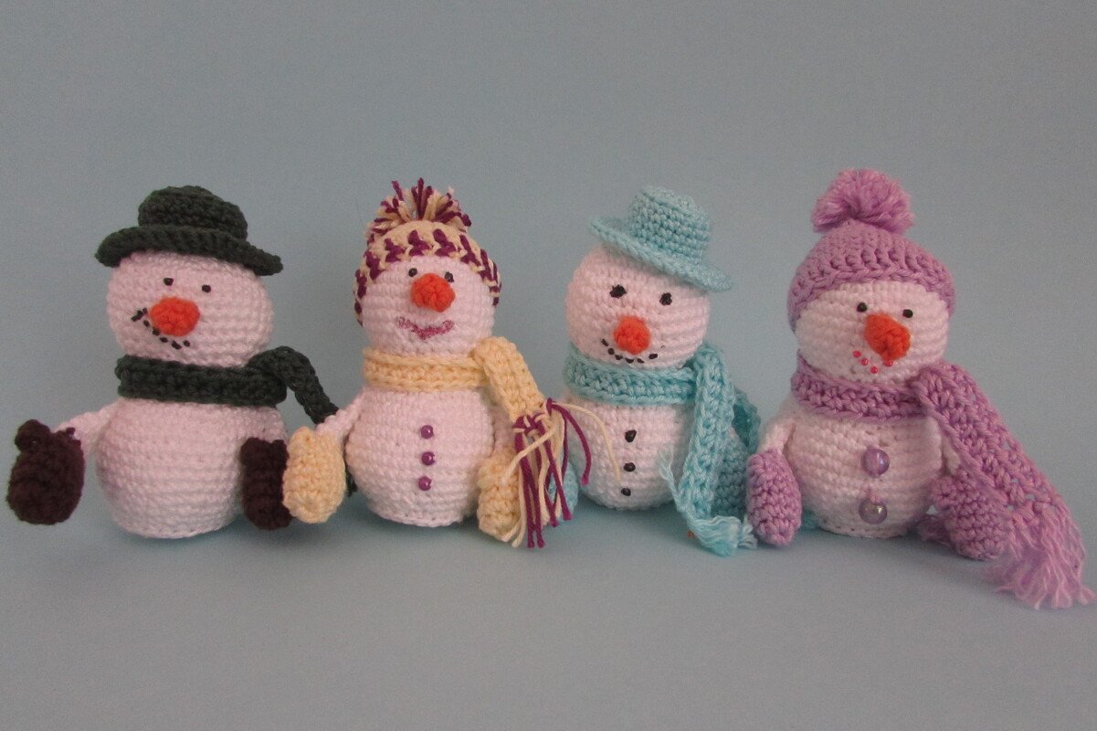 Vier gehäkelte Amigurumi‑Schneemänner mit bunten Mützen, Schals und Fäustlingen vor hellblauem Hintergrund.