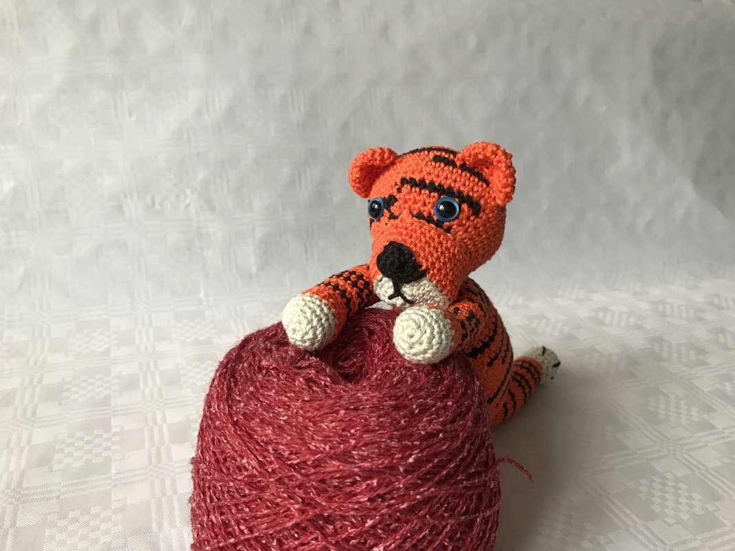 Kleiner orange-schwarz gestreifter Häkel-Tiger mit blauen Augen auf einem rötlichen Garnknäuel.