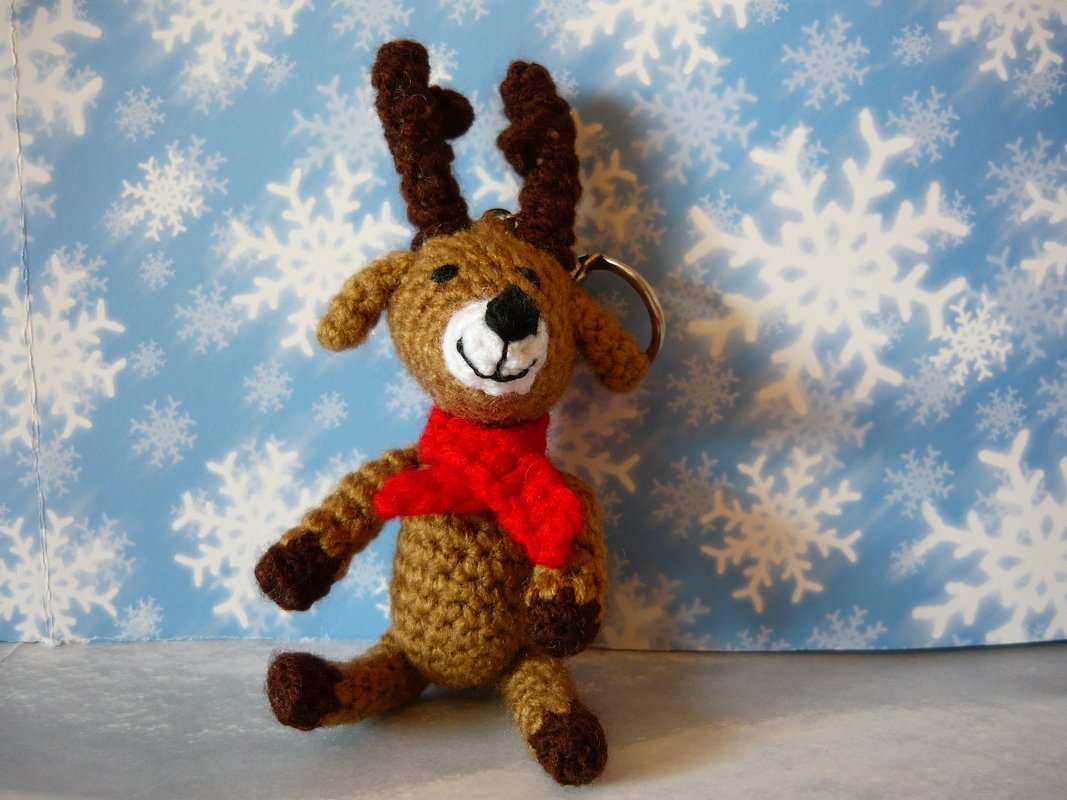 Gehäkeltes braunes Rentier-Amigurumi mit rotem Schal als Schlüsselanhänger vor Schneeflocken-Hintergrund