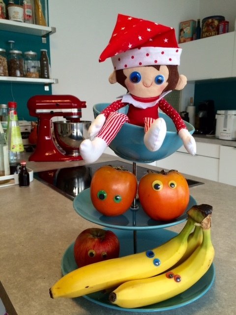 Genähte Wichtelpuppe auf dreistufiger Etagere über Bananen und Äpfeln mit bunten Knopfaugen