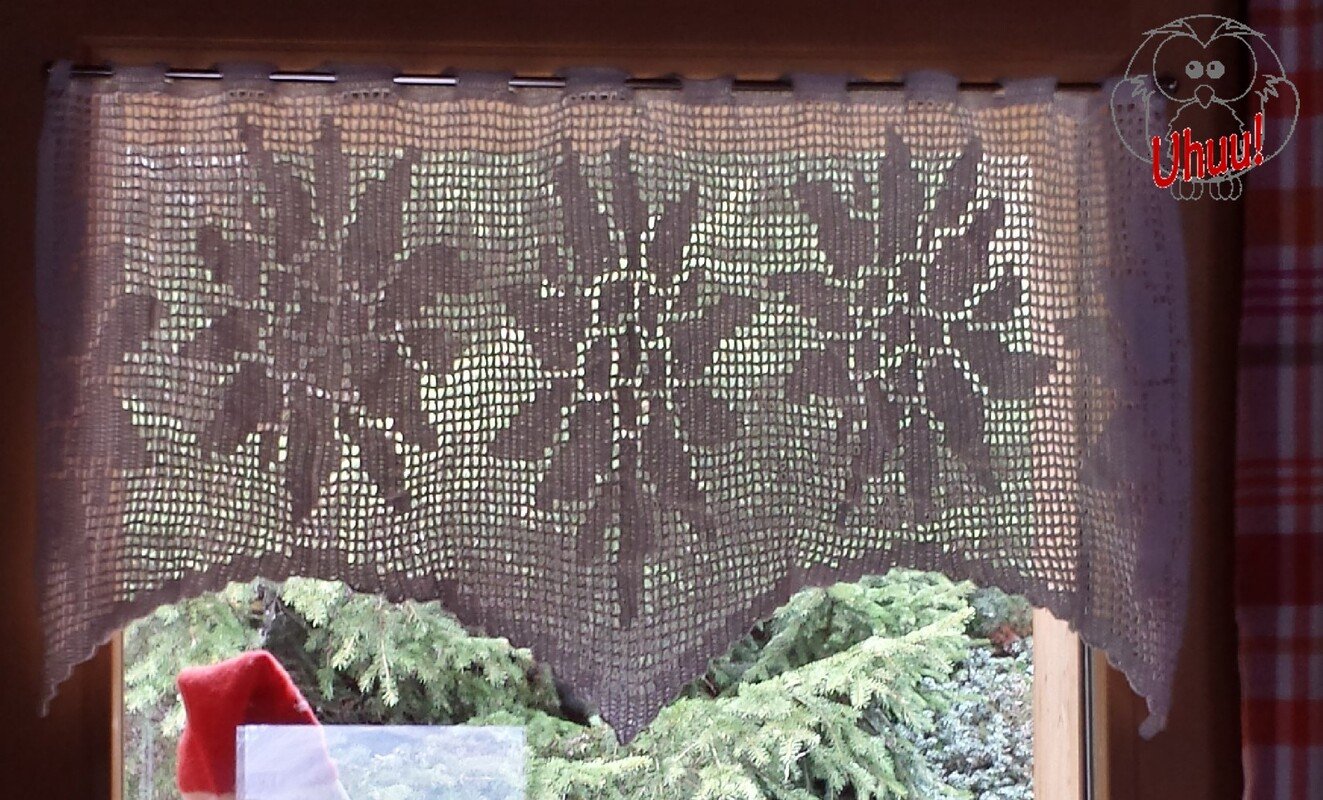 Weißer, gehäkelter Fensterbehang mit filigranen Schneeflockenmotiven vor einem Fenster, im Hintergrund grünes Gehölz