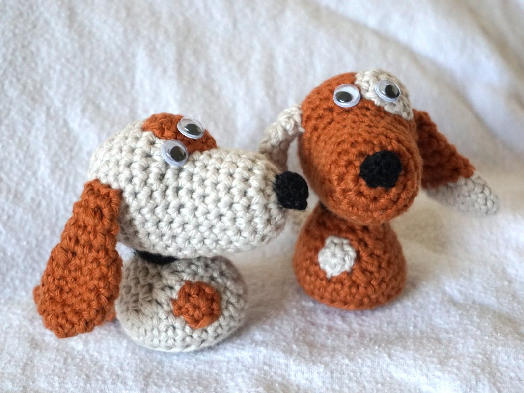 Zwei Amigurumi
Hunde, einer beige mit orangefarbenen Ohren und Punkten, der andere orange mit beigen Ohren, auf hellem Stoff.