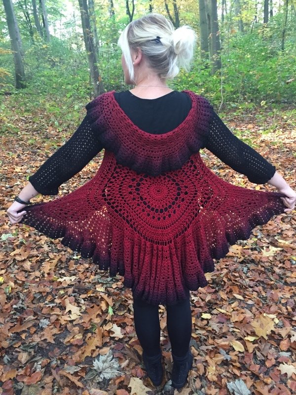 Rückenansicht einer Frau im Wald, trägt eine bordeauxrote bis schwarze gehäkelte Jacke mit rundem Mandala‑Motiv und weitem Saum