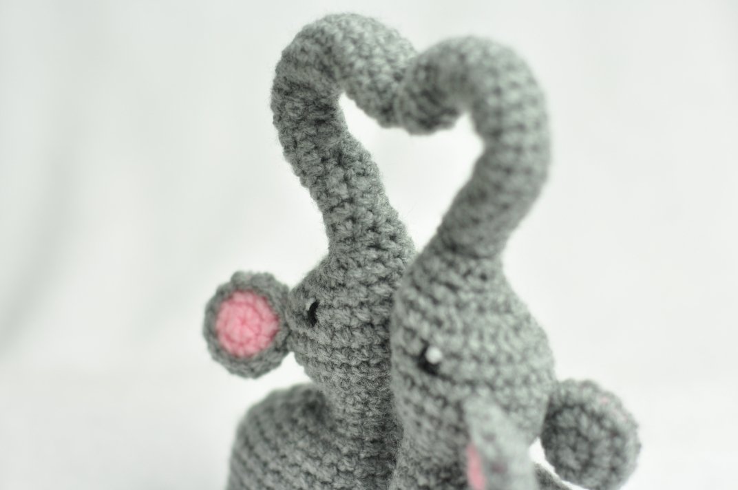 Grey amigurumi elephants with trunks forming a heart, showing stitch texture and pink ear detail