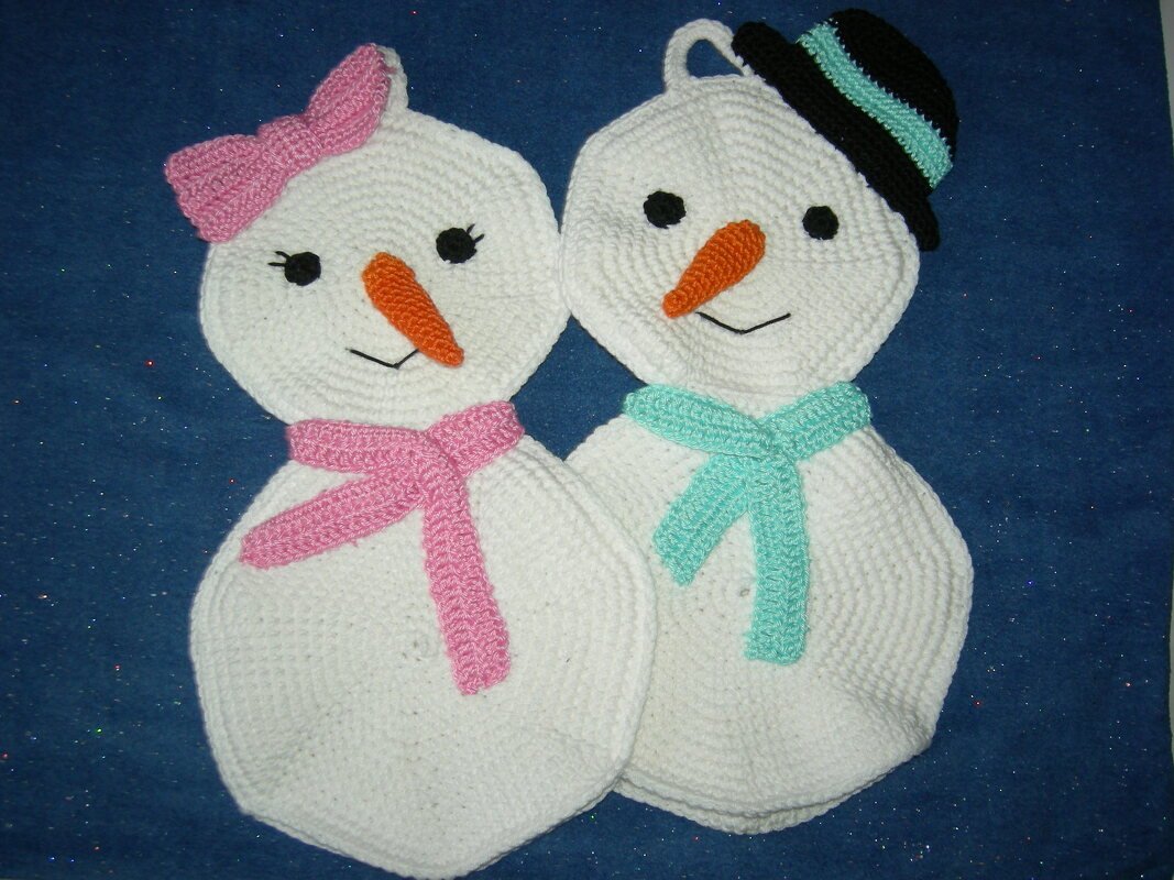 Zwei gehäkelte Schneemann-Topflappen auf blauem Hintergrund; links mit rosa Schleife und Schal, rechts mit schwarzem Hut und türkisfarbenem Schal.