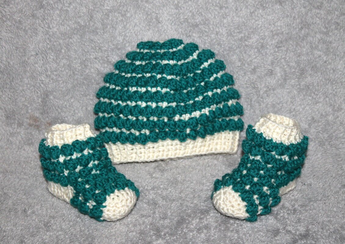 Gehäkeltes Baby-Set mit Mütze und zwei Schühchen in Petrol und Creme auf grauem Stoff.