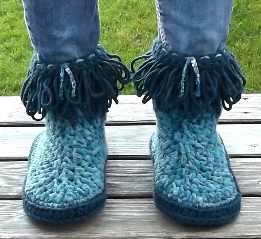 Paar gehäkelter türkiser Damenstiefel mit Fransen am Schaft, getragen zu Jeans auf Holzplanken.