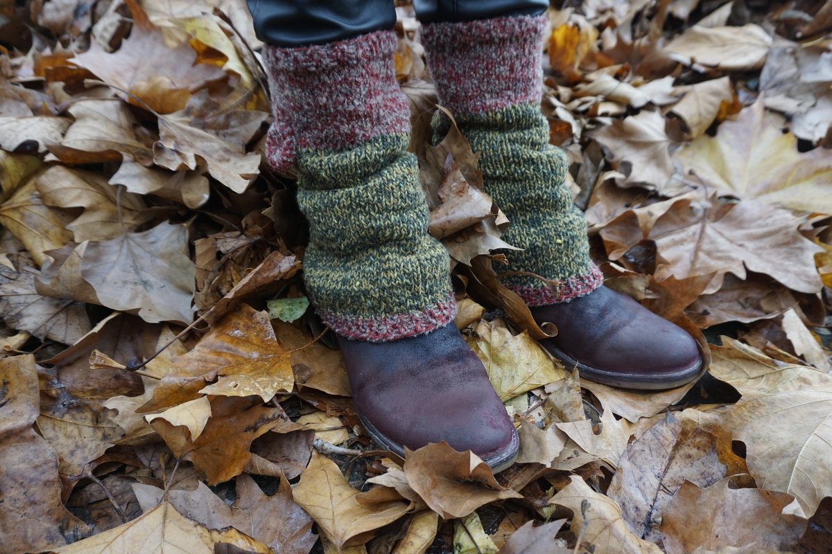 Zweifarbige, gestrickte Beinstulpen in Dunkelrot und Moosgrün über dunklen Schuhen auf trockenem Herbstlaub.
