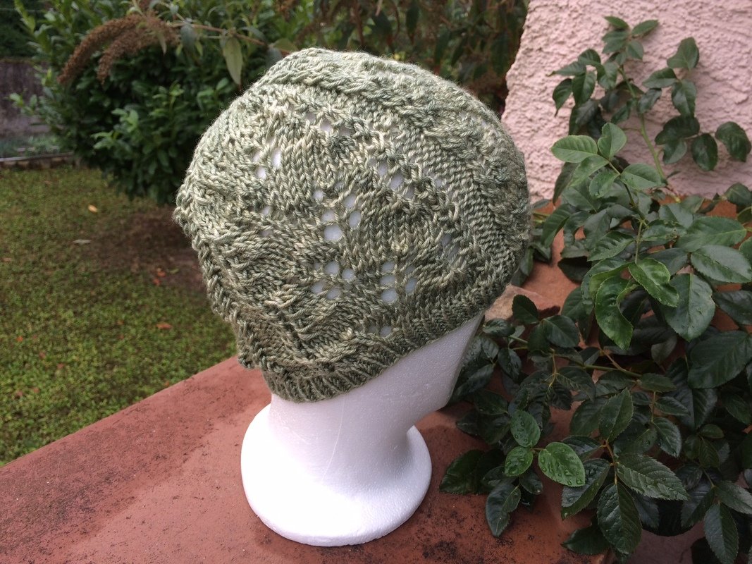 Salbeigrüne gestrickte Damenmütze mit Ajourmuster auf weißem Kopfmodell, umgeben von Pflanzen