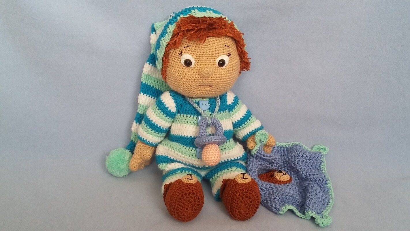 Hand-crocheted amigurumi doll in blue-green striped pajamas with sleeping cap, pacifier, and small animal-faced blanket