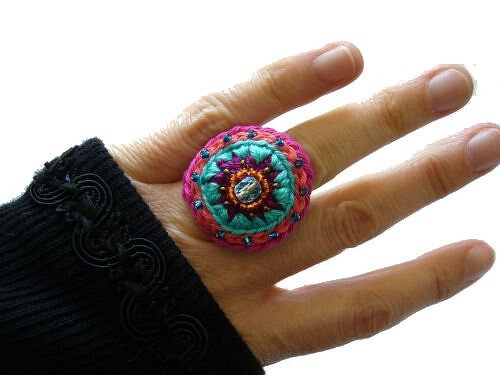 Hand mit schwarzem Ärmel trägt bunten runden Häkelring mit Perlen in Türkis, Pink und Orange