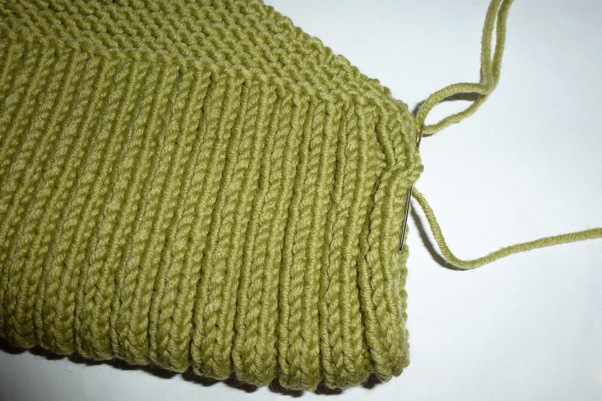 Nahaufnahme eines olivgrünen, gerippten Strickstücks mit Stopfnadel und losem Garn