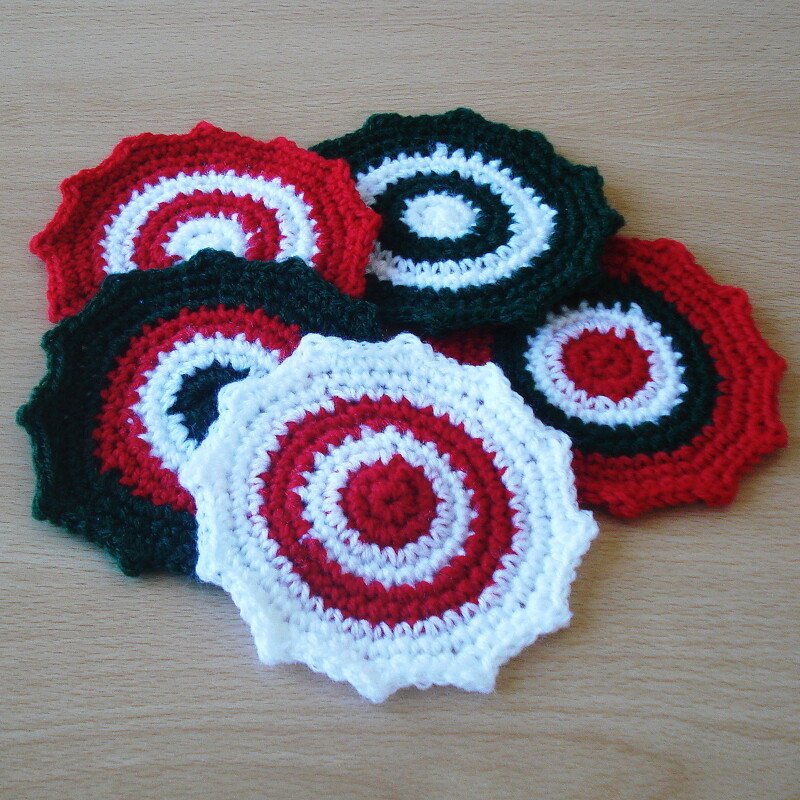 Five round crocheted coasters in red, white, and dark green with concentric rings and scalloped edges, stacked on a wooden surface.