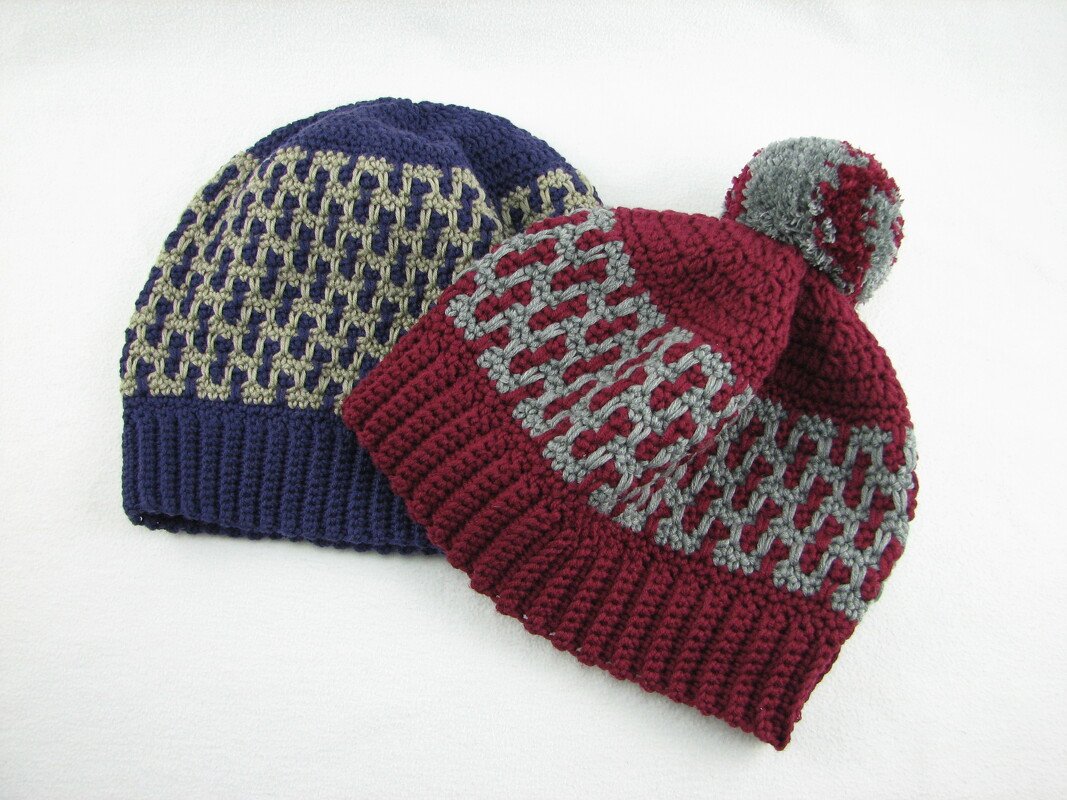 Zwei gehäkelte Mützen mit Gittermuster – dunkelblaue Beanie und bordeauxrote Bommelmütze.