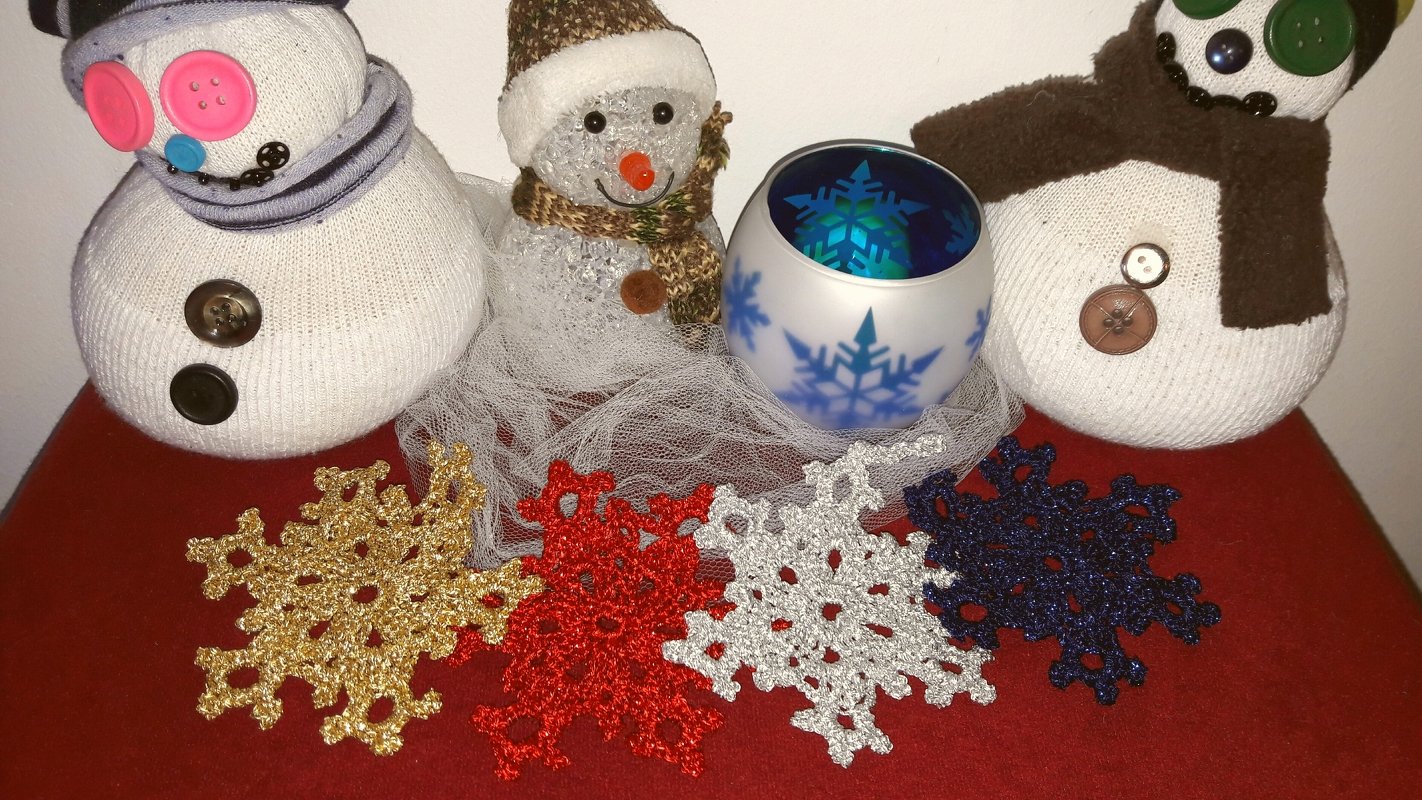 Drei Schneemänner, ein Schneeflocken-Teelichtglas und vier gehäkelte glitzernde Schneeflocken auf rotem Untergrund.