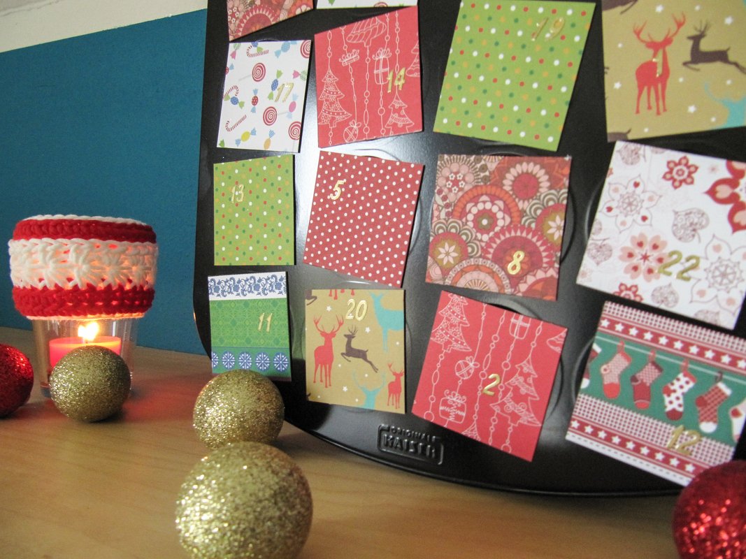 Adventskalender aus schwarzem Muffinblech mit nummerierten, gemusterten Papierklappen; daneben Kerze in gehäkelter Hülle und glitzernde Christbaumkugeln.