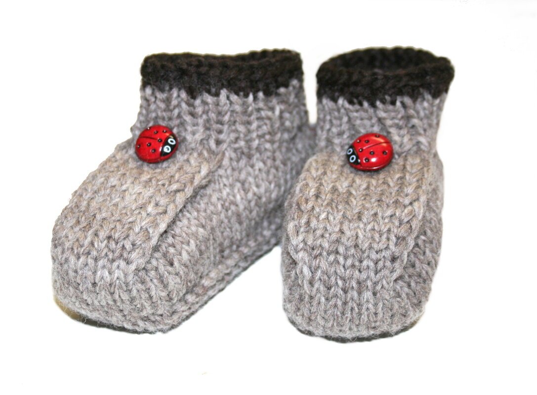 Hellgraue, handgestrickte Babyschuhe mit dunkelbraunen Bündchen und roten Marienkäfer-Knöpfen auf weißem Hintergrund.