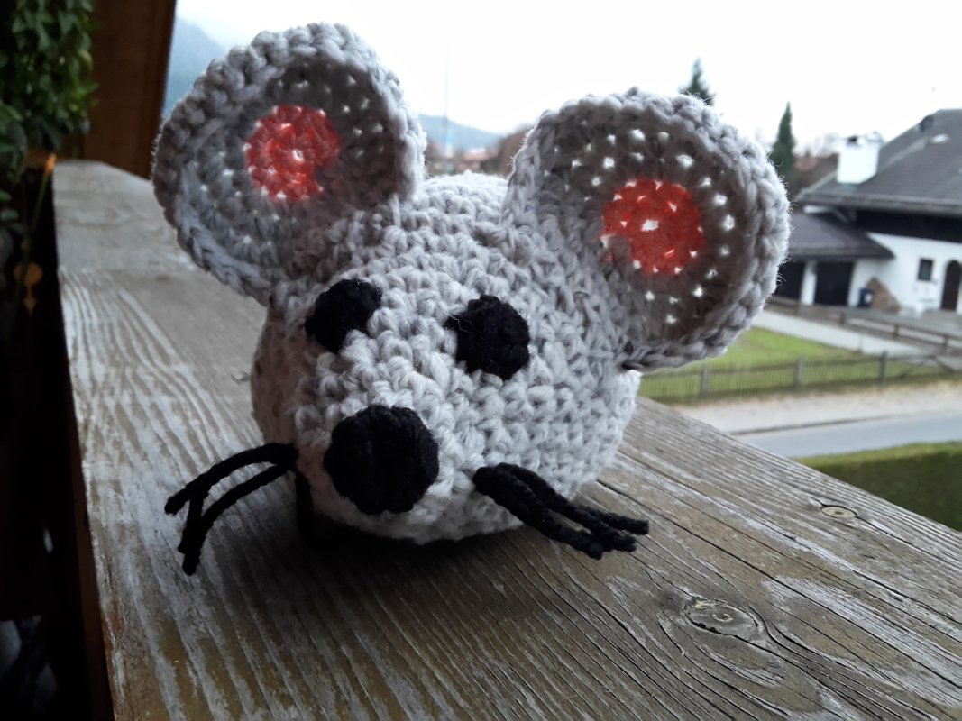 Gehäkelte graue Amigurumi-Maus mit orangefarbenen Innenohren, schwarzen Augen, Nase und Schnurrhaaren auf einem Holzbalken.