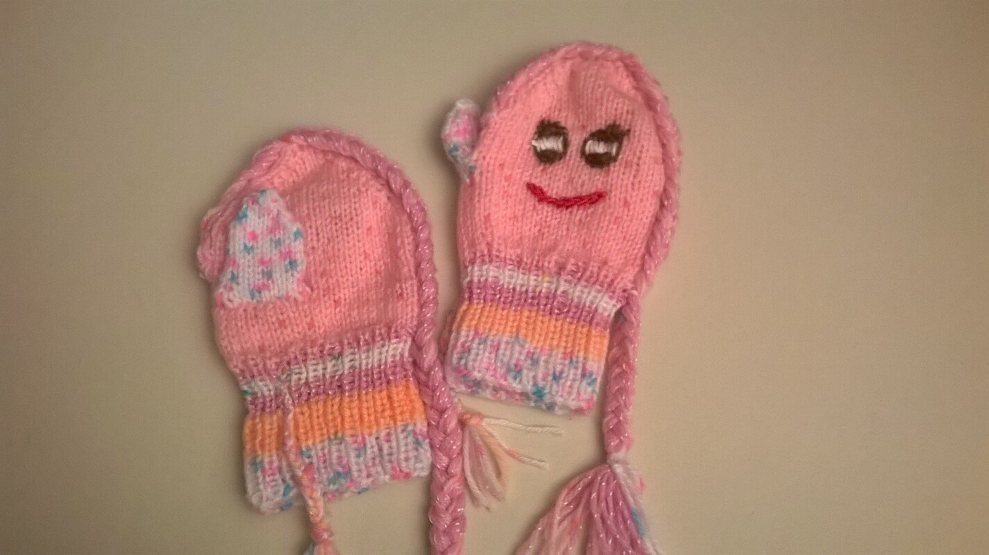 Gestrickte rosa Babyfäustlinge mit gesticktem Gesicht und geflochtenen Zöpfen