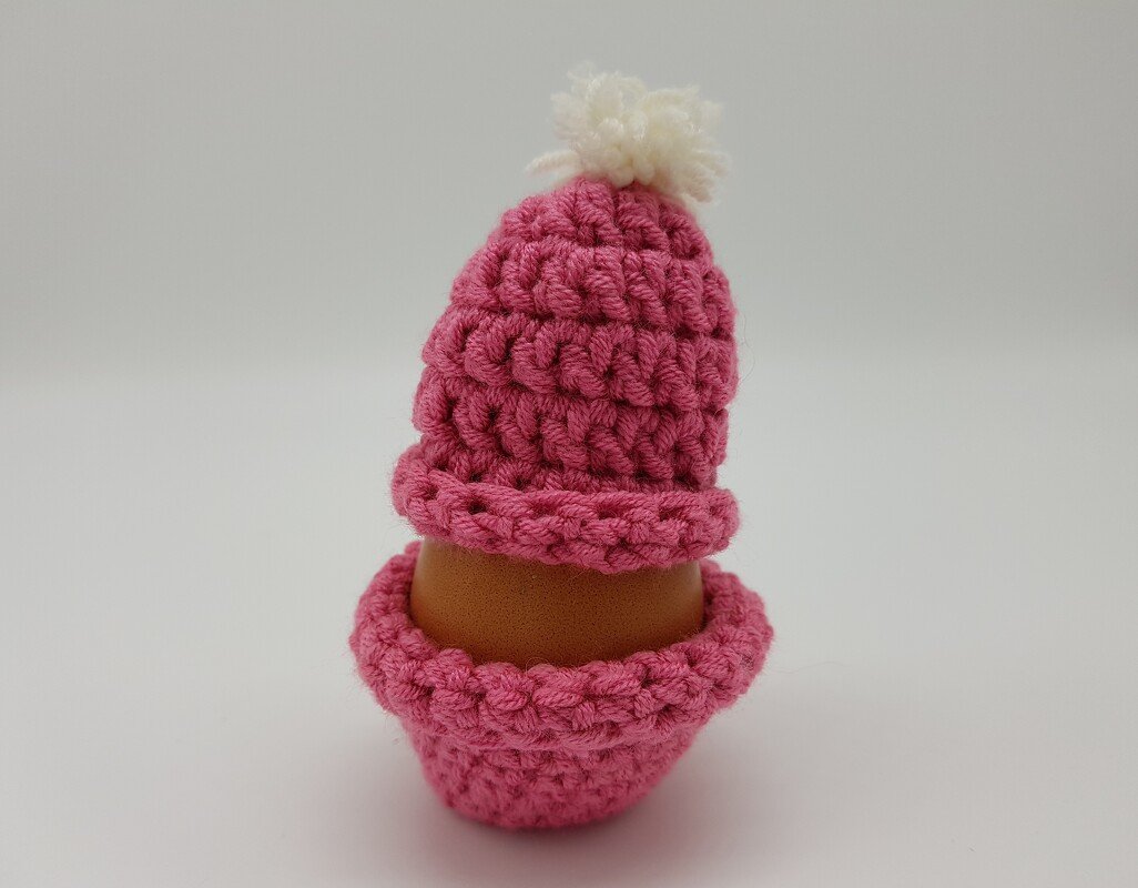 Braunes Ei im pinken gehäkelten Eierwärmer mit weißem Bommel vor hellem Hintergrund.