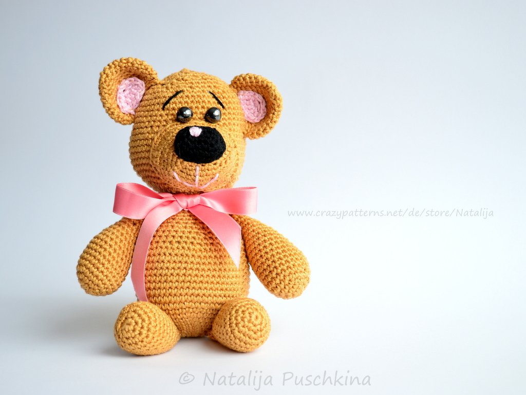 Sitzender gehäkelter Teddybär in Goldbraun mit rosa Schleifenband und rosa Ohren
