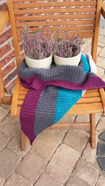 Gestricktes Tuch in Grau, Petrol und Violett auf Holzstuhl mit blühendem Heidekraut