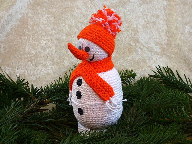 Gehäkelter Amigurumi-Schneemann mit orangefarbener Mütze und Schal auf Tannenzweigen
