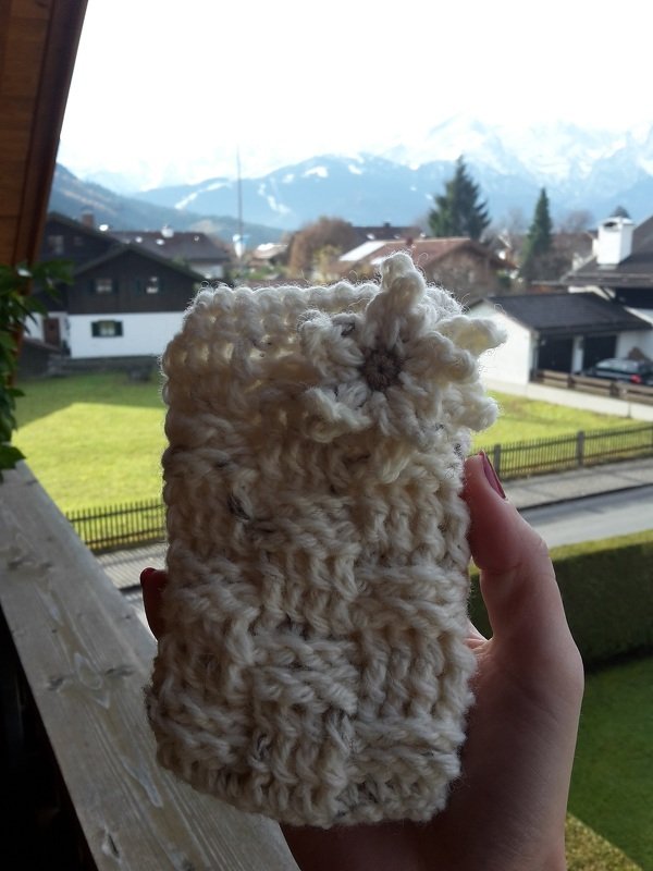 Nahaufnahme eines cremefarbenen gehäkelten Utensilos mit Zierblume, vor Häusern und Bergen gehalten.