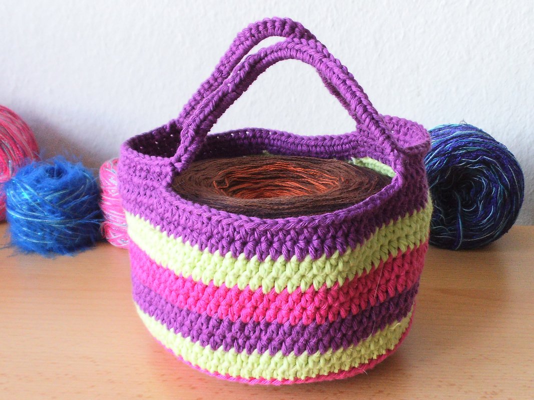 Rundes gehäkeltes Körbchen in lila, pink und gelb mit zwei Henkeln und Garnknäueln im Hintergrund