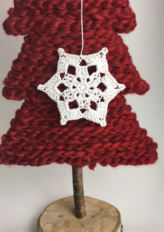 Weiße gehäkelte Schneeflocke an rotem, grob gestricktem Deko‑Tannenbaum mit Holzstamm