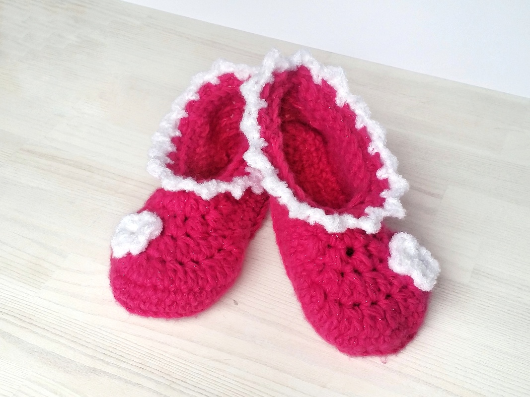 Zwei pinke gehäkelte Babyschuhe mit weißer Rüsche und kleinen Blüten auf hellem Holz