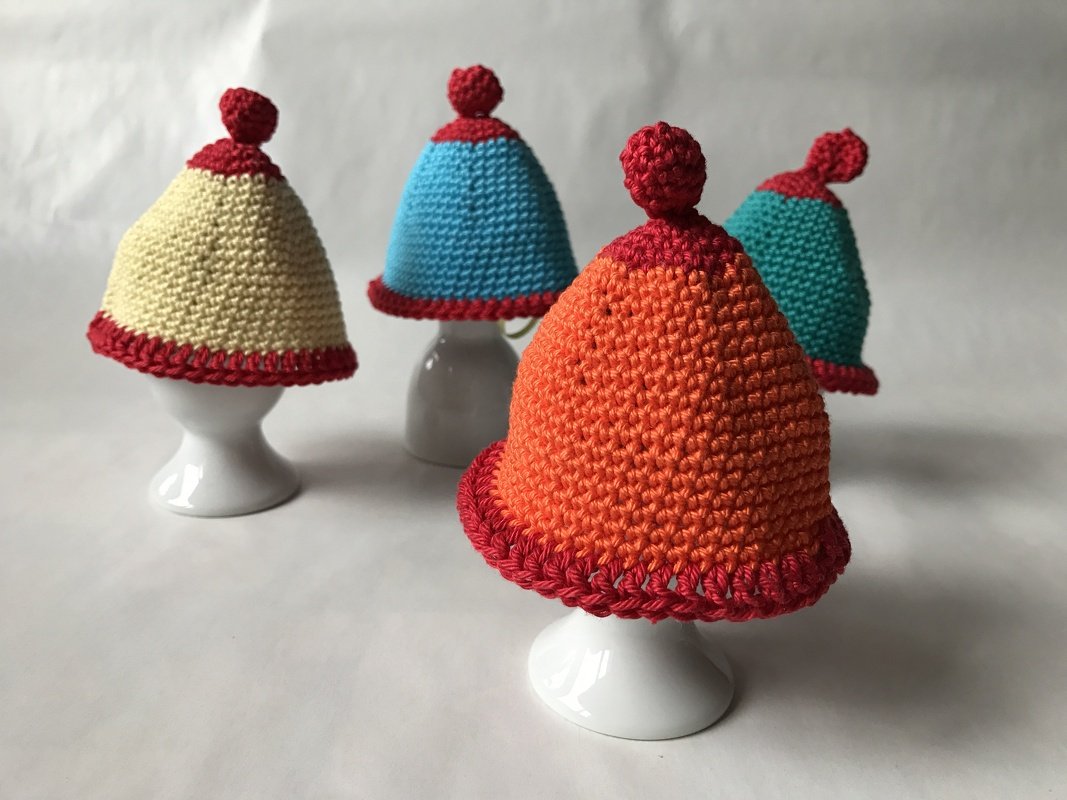 Vier gehäkelte Eierwärmer in Creme, Hellblau, Orange und Türkis mit rotem Rand und Bommel auf weißen Eierbechern vor hellem Hintergrund.