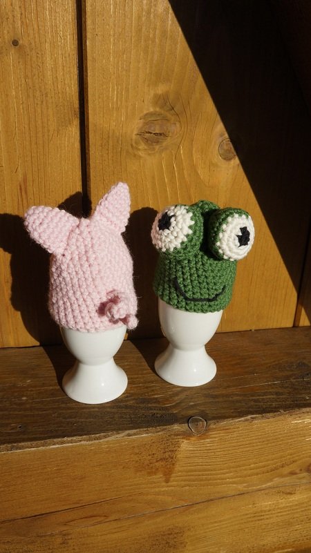 Rosa gehäkelter Hase und grüner Frosch als Eierwärmer in Eierbechern vor Holz