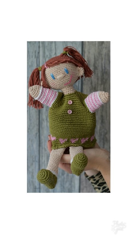 Gehäkelte Mädchen-Handpuppe mit olivgrünem Kleid, rosa Ärmeln und Zöpfen, gehalten von einer Hand