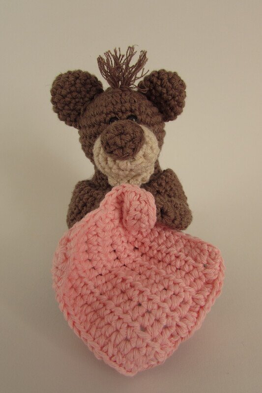 Nahaufnahme eines braunen Amigurumi-Teddybärs mit beiger Schnauze und fransiger Mähne, der ein rosafarbenes gehäkeltes Herz-Schnuffeltuch hält.