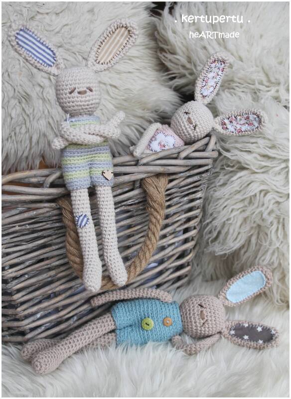 Drei gehäkelte Amigurumi-Hasen mit unterschiedlicher Kleidung auf hellem Fellteppich neben einem Weidenkorb.