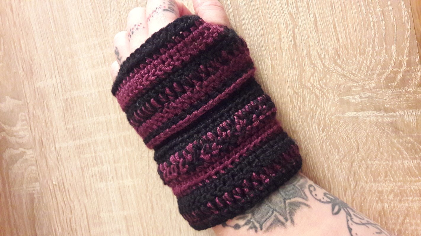 Hand mit gehäkelter Armstulpe in Schwarz und Weinrot auf Holzuntergrund