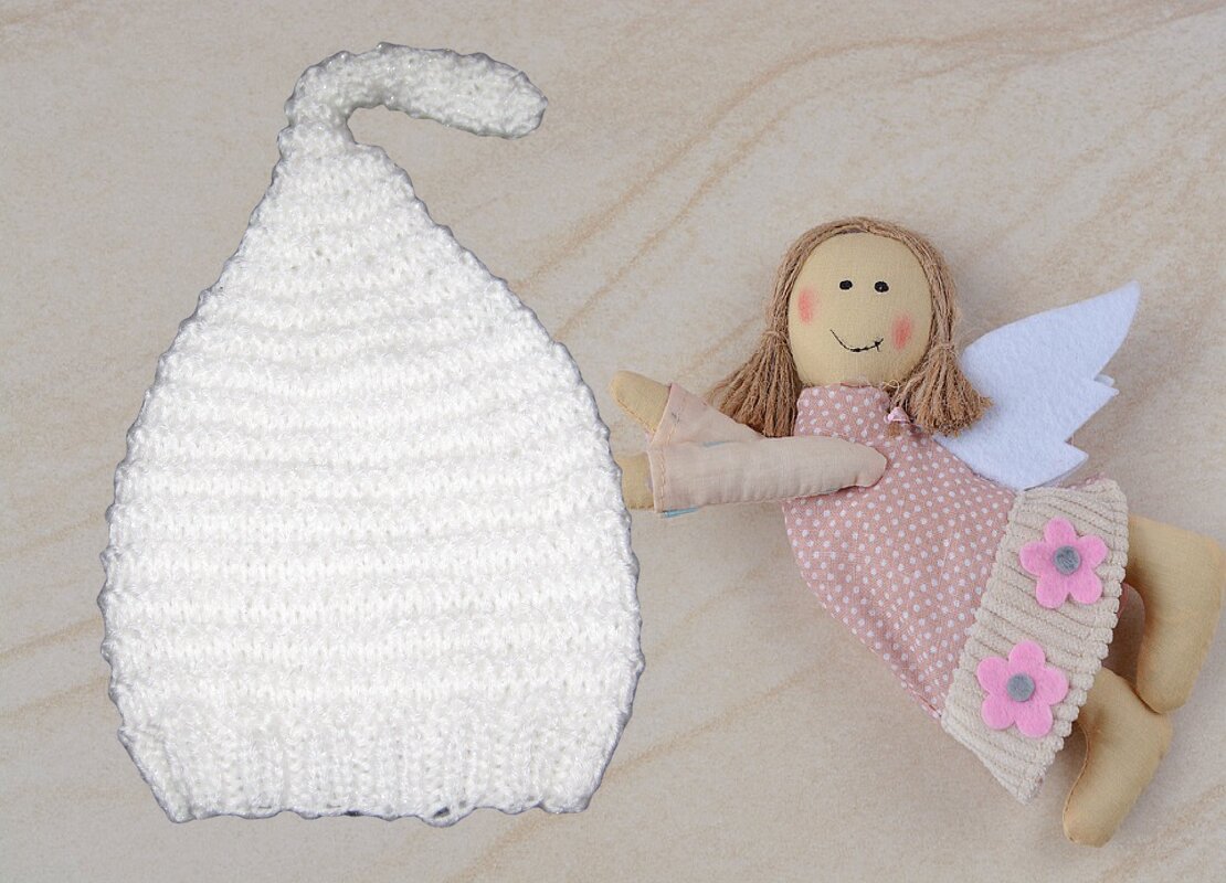 Weiße gestrickte Zipfel‑Babymütze neben einer Stoffpuppe